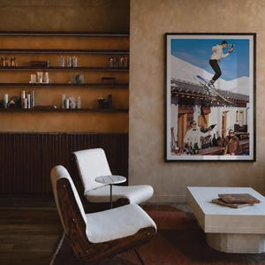 May include: A lounge area featuring two white upholstered chairs with dark wood frames, a small round side table, and a low white rectangular coffee table. A large framed photograph on the wall depicts a skier jumping over a building and a bar scene.