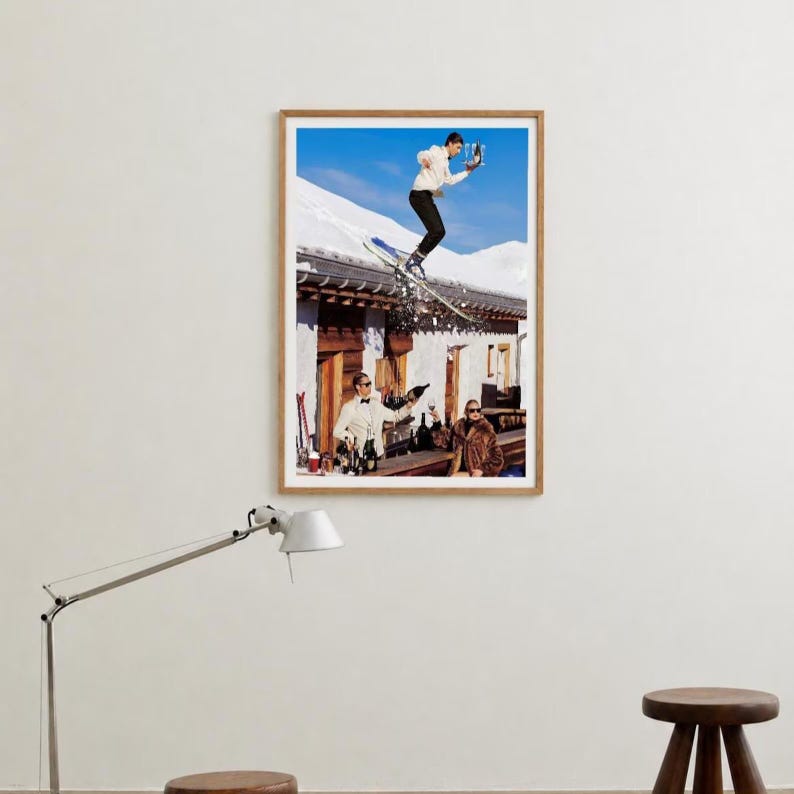 May include: A framed wall art print depicting a skier in a white jacket jumping over a rustic wooden building with a snow-covered roof. Below, two individuals in formal attire stand near a bar counter, one holding a bottle and the other a glass.