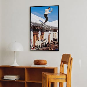 May include: A framed wall art print featuring a surreal scene of a person in a white sweater and black pants jumping on skis over a building roof while holding a tray of drinks, with two people below at a bar counter against a snowy mountain backdrop.