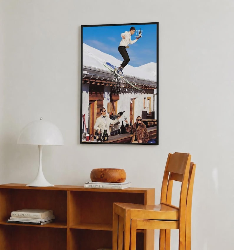 May include: A framed wall art print featuring a surreal scene of a person in a white sweater and black pants jumping on skis over a building roof while holding a tray of drinks, with two people below at a bar counter against a snowy mountain backdrop.