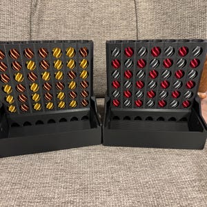 May include: Two black Connect Four game boards. One board has red, silver, and black game pieces. The other board has gold, orange, and brown game pieces. The boards are sitting on a gray upholstered chair.