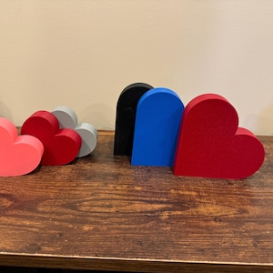 Puede incluir: Cinco adornos de madera en forma de corazón de diferentes colores. Los corazones son rosa, rojo, gris, negro y azul.
