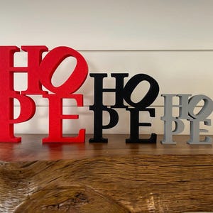 Puede incluir: Tres letreros de madera que deletrean "HOPE" en diferentes tamaños. El letrero más grande es rojo, el letrero del medio es negro y el letrero más pequeño es gris.