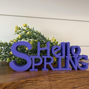 May include: Purple wooden sign with the words "Hello Spring" in a decorative font. The sign is sitting on a wooden surface with a bouquet of yellow flowers in the background.