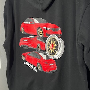 Peut inclure: Sweat-shirt noir avec un motif de voiture rouge au dos. La voiture est une BMW M3 et le texte "DRVN" et "DRIVEN.EU" sont imprim&eacute;s sur le sweat-shirt.