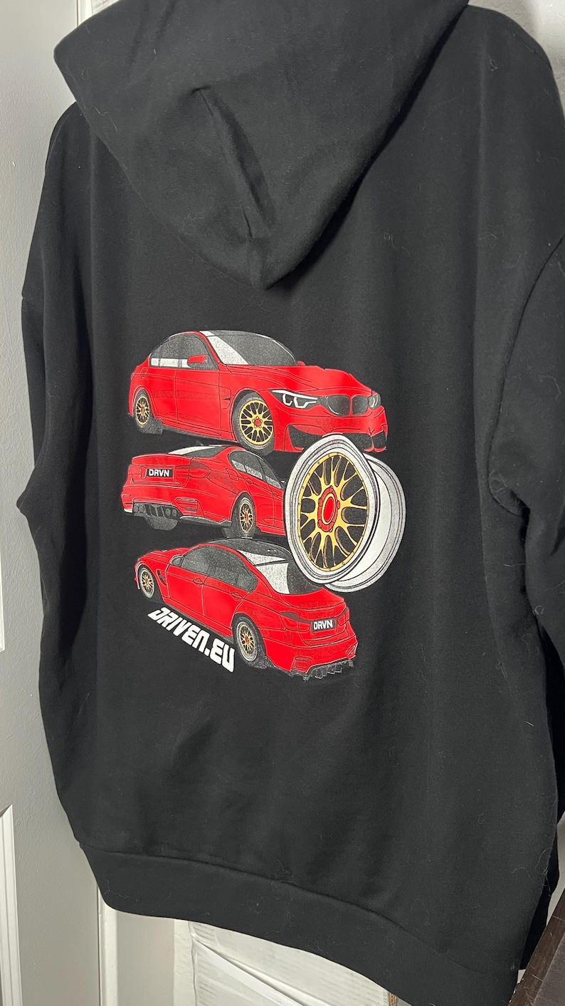 Peut inclure: Sweat-shirt noir avec un motif de voiture rouge au dos. La voiture est une BMW M3 et le texte "DRVN" et "DRIVEN.EU" sont imprim&eacute;s sur le sweat-shirt.
