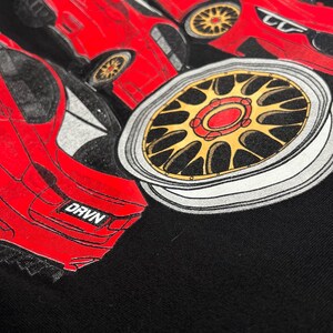 Peut inclure: Un sweat-shirt noir avec un imprim&eacute; graphique de voiture rouge. La voiture a une jante dor&eacute;e et le texte "DRVN" est visible &agrave; l'arri&egrave;re de la voiture.