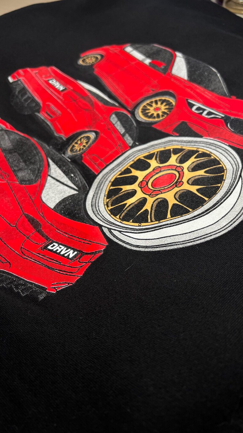 Peut inclure: Un sweat-shirt noir avec un imprim&eacute; graphique de voiture rouge. La voiture a une jante dor&eacute;e et le texte "DRVN" est visible &agrave; l'arri&egrave;re de la voiture.