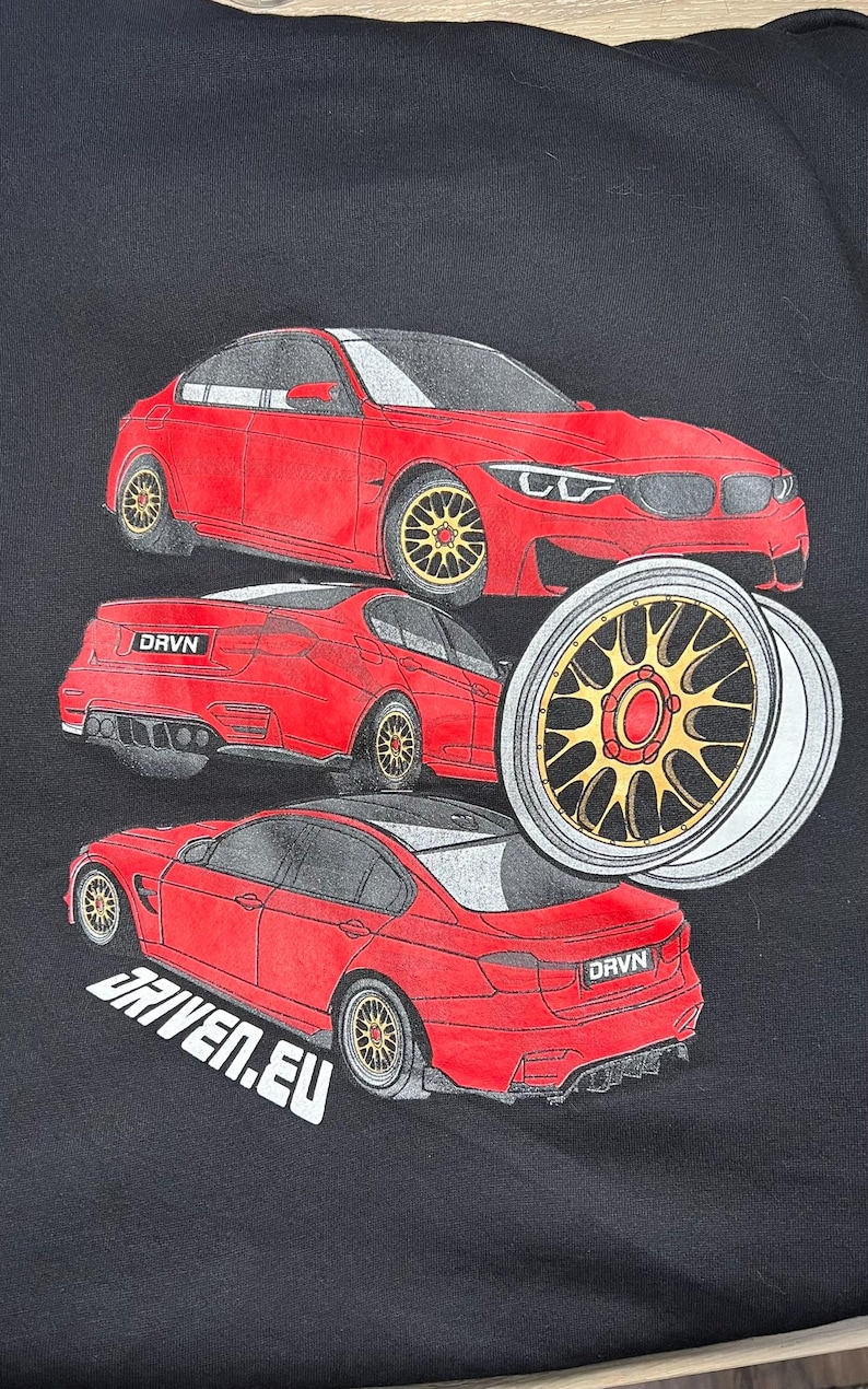 Peut inclure: Un t-shirt noir avec un motif de voiture rouge. La voiture est repr&eacute;sent&eacute;e sous diff&eacute;rents angles, notamment de face, de dos et de c&ocirc;t&eacute;. La voiture a des roues noires avec des accents dor&eacute;s. Le texte "DRVN" et "DRIVEN.EU" est imprim&eacute; sur le t-shirt.
