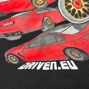 Peut inclure: Un t-shirt noir avec un motif rouge, blanc et gris de trois voitures. Les voitures sont toutes rouges et ont des roues noires. Le texte "DRVN" et "DRIVEN.EU" est imprim&eacute; sur le t-shirt.