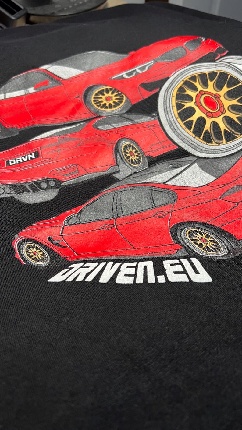 Peut inclure: Un t-shirt noir avec un motif rouge, blanc et gris de trois voitures. Les voitures sont toutes rouges et ont des roues noires. Le texte "DRVN" et "DRIVEN.EU" est imprim&eacute; sur le t-shirt.