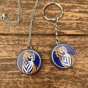 May include: A silver-toned metal necklace with a circular pendant and a matching circular keyring, both featuring a graphic of a roaring tiger in orange, white, and blue colours set against a dark blue background.