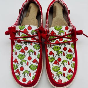 Puede incluir: Un par de zapatos rojos sin cordones con un interior blanco con un estampado del Grinch. El Grinch lleva un gorro de Papá Noel y sostiene un adorno rojo. Los zapatos tienen cordones rojos y un ribete rojo.
