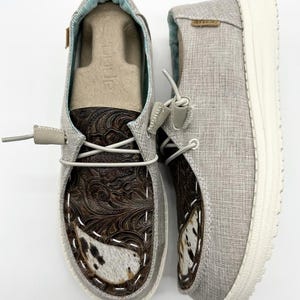 May include: A pair of gray canvas slip-on shoes with brown leather and white cowhide accents. The shoes have a white sole and a lace-up design.