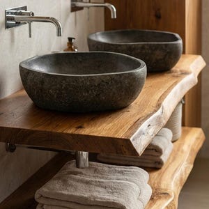 Peut inclure: Meuble-vasque de salle de bain avec deux vasques en pierre gris foncé posées sur une étagère en bois aux bords naturels. L'étagère est soutenue par des supports métalliques. Des serviettes beiges sont empilées en dessous.