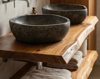 Könnte beinhalten: Zwei dunkelgraue Steinwaschbecken sind auf einer Waschtischplatte aus Naturholz mit Baumkante montiert. Der Waschtisch hat zwei Ebenen, wobei gefaltete beige Handtücher auf der unteren Ablage liegen. Zwei verchromte Wandarmaturen sind über den Becken.