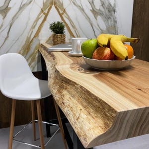Peut inclure: Une table en bois avec un bord naturel, ornée d'un bol de fruits : bananes, pommes et une orange. Une chaise blanche avec des pieds en bois est à proximité. Une petite plante en pot et un ordinateur portable sont également visibles.