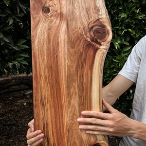 Puede incluir: Una tabla de cortar de madera grande y rectangular con un patrón de veta de madera natural. La tabla tiene una forma única e irregular con un nudo en un lado. La tabla está hecha por R&R Wood Studio.