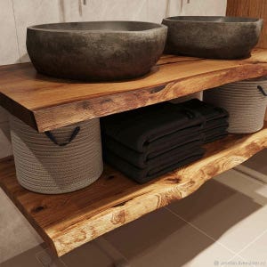 Peut inclure: Meuble-lavabo de salle de bain avec deux vasques en pierre gris foncé sur une étagère en bois naturel. L'étagère a un bord brut. En dessous, des serviettes noires pliées et deux paniers tressés avec des poignées noires. Dans une salle de bain moderne.