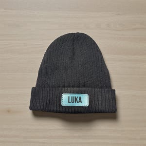 May include: A dark gray ribbed knit beanie with a folded brim. A light blue rectangular patch with the name "LUKA" in white letters is sewn onto the front of the hat. The hat is displayed on a light wood surface.