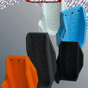 Könnte beinhalten: Eine Sammlung von fünf Beyblade Customs Launcher-Haltern aus Kunststoff in Orange, Schwarz, Hellblau und Weiß. Jeder Halter hat eine vertikale Ausrichtung mit einem vertieften Mittelkanal, drei kleinen kreisförmigen Löchern an der Oberseite und dem geprägten Text Beyblade Customs.