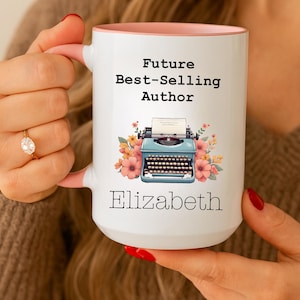 May include: White ceramic mug with a pink interior and handle, featuring the text "Future Best-Selling Author" above a vintage blue typewriter illustration and the name "Elizabeth". Floral accents surround the typewriter.