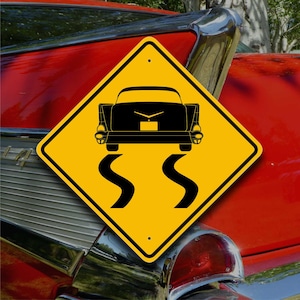 May include: Yellow diamond-shaped road sign with a black silhouette of a classic car driving over a winding road. The sign reads "S S" in black.