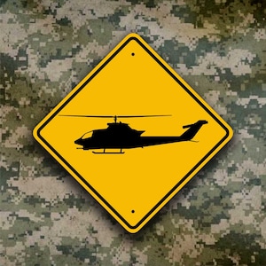 May include: Yellow diamond-shaped road sign with a black silhouette of a helicopter. The sign has a black border and a white dot in each corner.