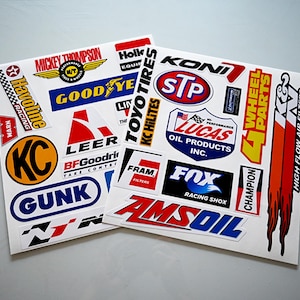 May include: A sheet of white paper with various colorful decals featuring brand names like Havoline, Good Year, Koni, STP, Lucas Oil Products, Champion, and more. The decals are for automotive and racing purposes.