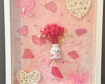 Könnte beinhalten: Gerahmtes Kunstwerk in Rosa und Weiß. Es zeigt rosa Schmetterlinge, geflochtene Herzen, rosa Trockenblumen in einer kleinen Vase und rosa Papierakzente. Der weiße Rahmen umschließt die Komposition.