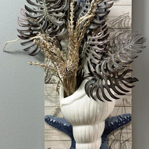 May include: Coastal-themed wall decor featuring a white seashell vase filled with dried plants and dark gray leaves. A blue starfish and seashell accents are mounted on a wooden plank with nautical illustrations.