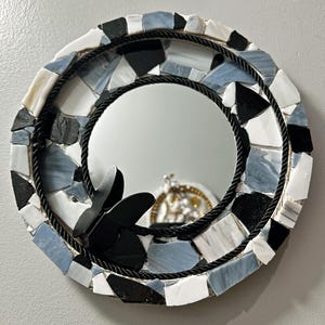 Könnte beinhalten: Runder Mosaikspiegel mit schwarzem Seilrand und einem schwarzen Schmetterlingsakzent. Das Mosaik zeigt eine Mischung aus weißen, schwarzen und hellblauen Fliesen. Der Spiegel reflektiert ein kleines dekoratives Objekt.
