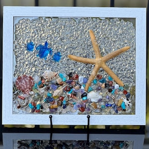 以下が含まれることがあります： ヒトデ、貝殻、カラフルな石が埋め込まれた樹脂製の額装アート。背景はテクスチャのある透明な樹脂です。
