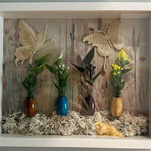 May include: A framed shadow box featuring two embroidered butterflies and miniature floral arrangements in small vases. The background has a rustic, wood-like appearance, and the base is covered in dried moss. The frame is white.