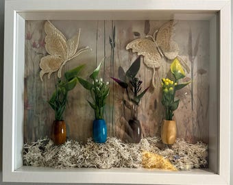 Könnte beinhalten: Ein gerahmter Schattenkasten mit zwei gestickten Schmetterlingen und Miniatur-Blumenarrangements in kleinen Vasen. Der Hintergrund hat ein rustikales, holzähnliches Aussehen, und die Basis ist mit getrocknetem Moos bedeckt. Der Rahmen ist weiß.