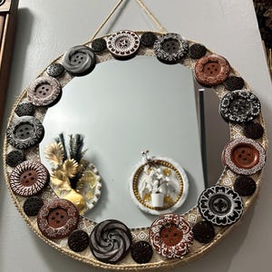 May include: Round mirror with a decorative border of various buttons in shades of brown, black, and white. The mirror is framed with a woven jute rope and a patterned fabric. A natural fiber cord is used for hanging.