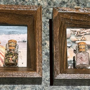 以下が含まれることがあります： 2つの額装されたアートワークは、それぞれコルク栓付きの小さなガラス瓶を3つずつ特徴としています。ボトルには様々なアイテムが入っており、ビーチの風景とテクスチャード加工の背景が背景にあります。フレームは濃い色の木材で作られています。