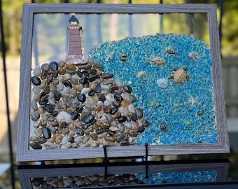 May include: A framed mosaic artwork depicting a lighthouse on a rocky shore with blue glass representing the ocean. The mosaic includes seashells, pebbles, and a starfish.