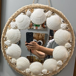 May include: Round mirror with a decorative border of rope and seashells. The mirror's frame is adorned with large, white seashells and smaller shells, creating a coastal theme. A natural rope is used for hanging.