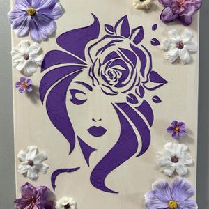 May include: A canvas artwork depicting a purple silhouette of a woman's face and hair, with a rose detail. The piece is decorated with artificial flowers in purple and white, giving a three-dimensional appearance.
