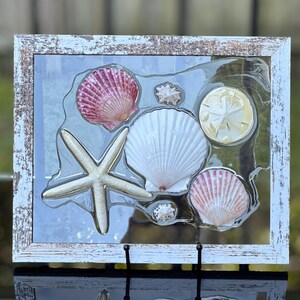 May include: A framed seashell art piece with a white frame and a clear glass insert. The piece features a variety of seashells, including a large white scallop shell, a pink and white scallop shell, a sand dollar, and a starfish.