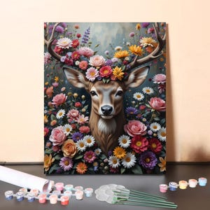 Puede incluir: Un kit de pintura por números con un ciervo con una corona floral. El ciervo está rodeado de flores coloridas en tonos rosa, amarillo, naranja y morado. El kit incluye botes de pintura, pinceles y un lienzo.