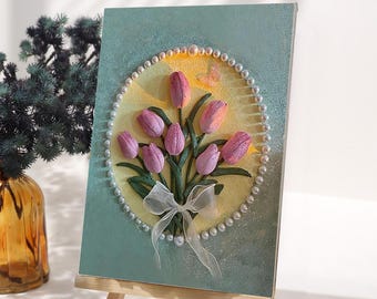 May include: A framed artwork featuring a bouquet of pink tulips, accented with green leaves and a white ribbon. The tulips are arranged in a circle, surrounded by a pearl border, set against a light blue and yellow background.