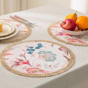 May include: Circular floral placemats with a cream backdrop, adorned with pink, blue, and green floral designs. Each mat has a decorative gold trim. The placemats are arranged on a white tablecloth, alongside a plate, cutlery, and a fruit bowl.