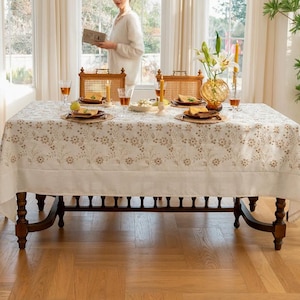 May include: A rectangular dining table covered with a white tablecloth featuring an embroidered tan floral pattern along the edges. The table is set with woven placemats, ceramic plates, glassware, candles, and a glass vase with white lilies.