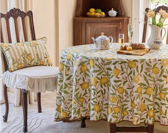 May include: A dining room scene featuring a round table covered with a lemon-themed tablecloth. A matching pillow rests on a chair. The tablecloth and pillow feature a pattern of yellow lemons and green leaves. A teapot and a vase with flowers are on the table.