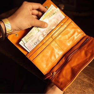 Könnte beinhalten: Eine offene, orangefarbene Ledergeldbörse mit mehreren Fächern, darunter ein Reißverschlussfach. Die Geldbörse ist mit verschiedenen Banknoten gefüllt. Eine goldene Uhr und ein schwarzes Armband sind sichtbar.