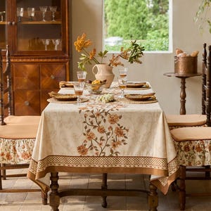May include: A dining table set for a meal. The table is covered with a cream-colored tablecloth with a floral pattern and brown border. Wooden chairs with floral seat covers surround the table. A vase of flowers and glasses are on the table.