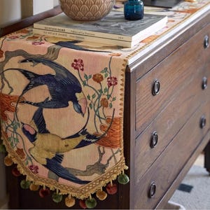 May include: A decorative table runner featuring a design of swallows in flight, with floral and branch accents. The runner has a pink background with a gold trim and pom-pom fringe. The runner is placed on a wooden dresser.