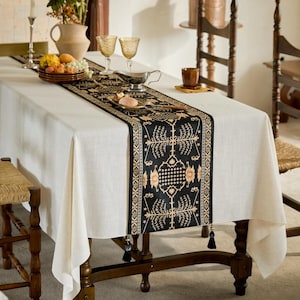 Puede incluir: Una mesa de comedor con un mantel blanco y un camino de mesa con estampado negro y dorado. La mesa está puesta con cristalería, una salsera y un plato de fruta. El camino de mesa tiene borlas en cada extremo.